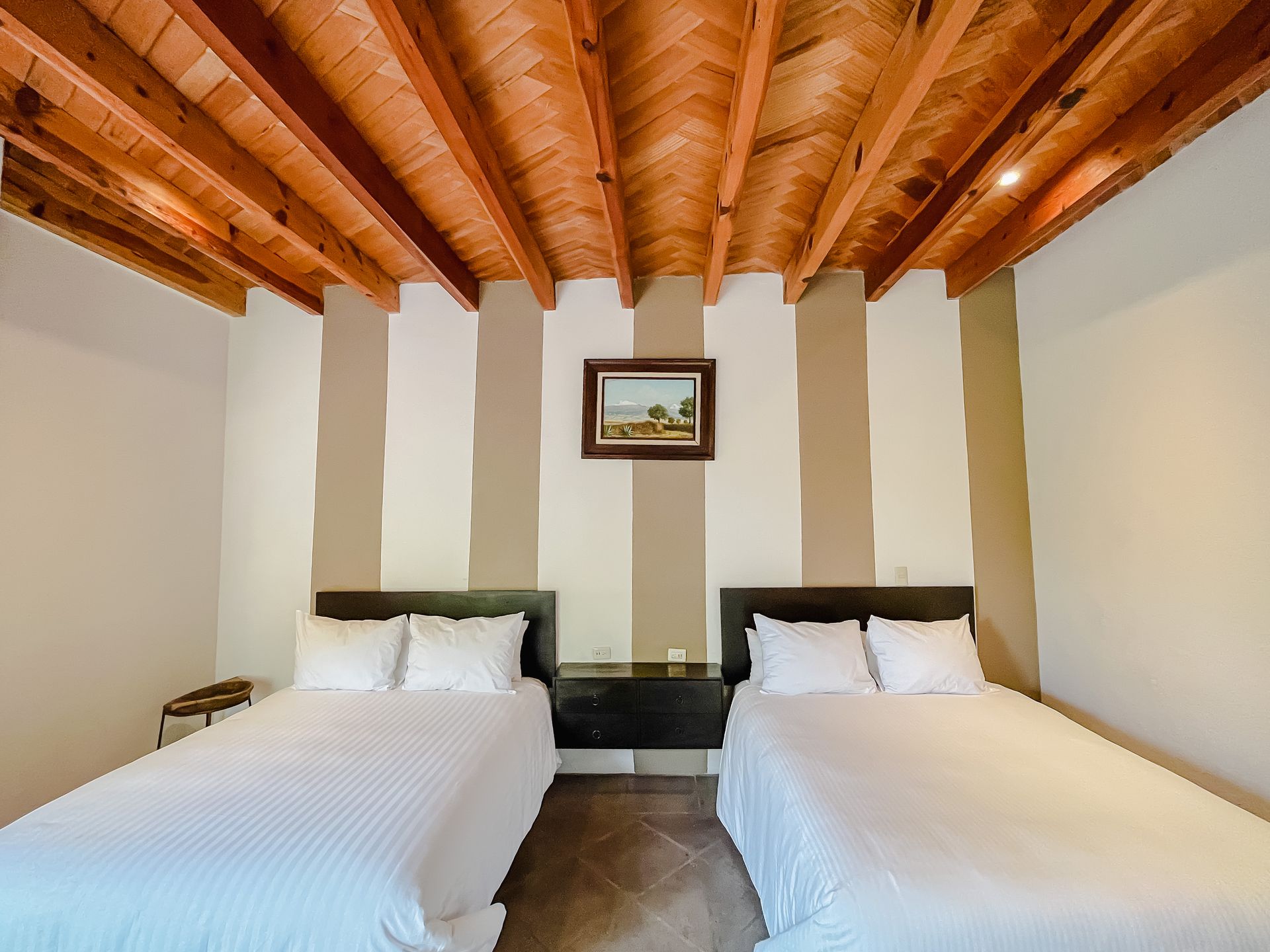 Dos camas en una habitación con techo de madera, paredes rayadas y un pequeño cuadro enmarcado.