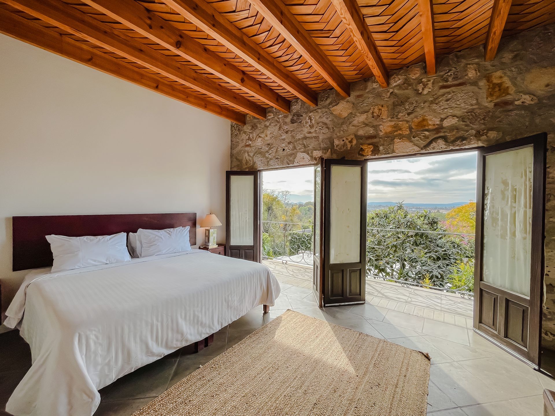 Dormitorio con cama, paredes de piedra, puertas abiertas y vista a los árboles y al cielo.