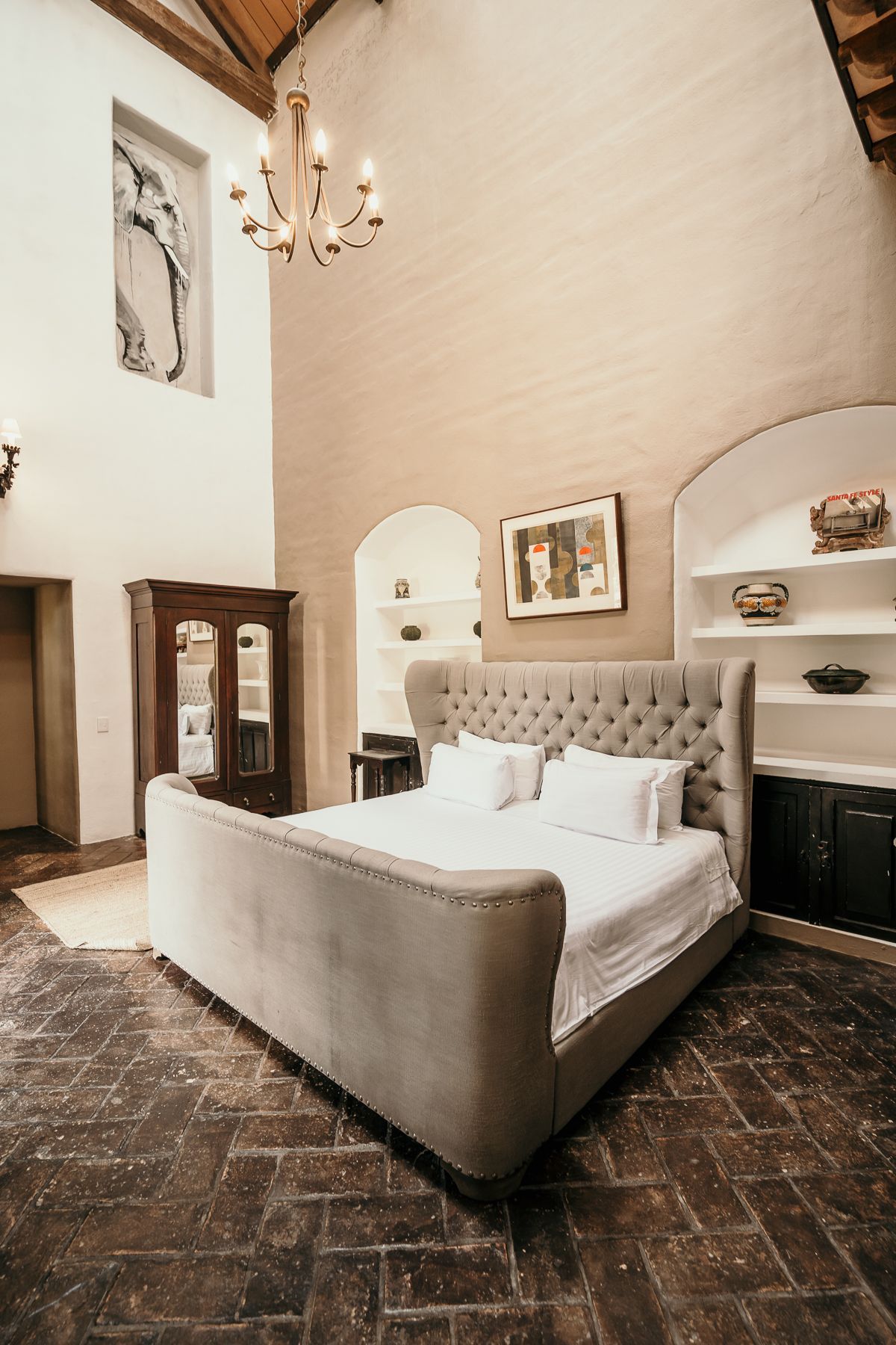 Dormitorio con cama capitoné gris, techos altos, lámpara de araña, estantes arqueados y suelo de ladrillo oscuro.