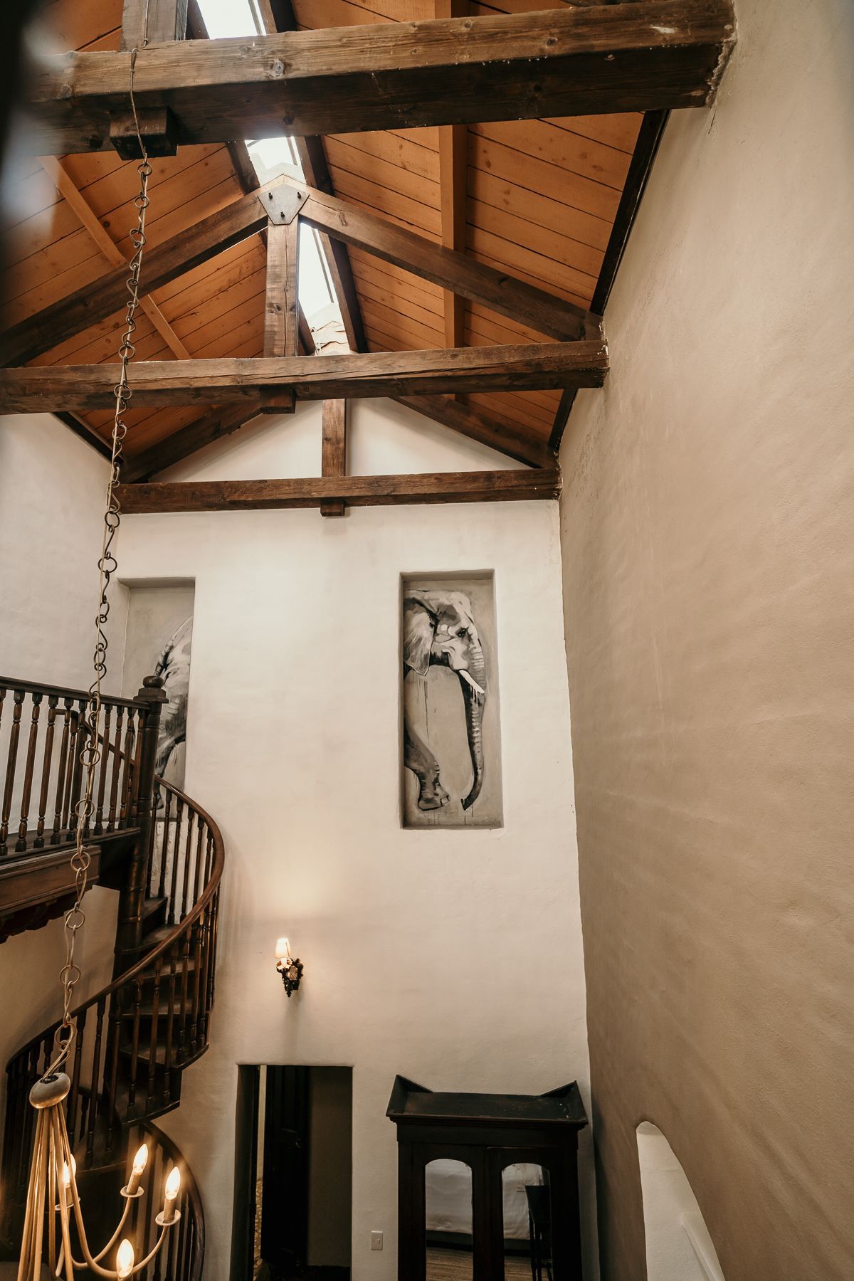 Escalera de caracol de madera y techo alto con vigas a la vista, obras de arte en la pared y lámpara de araña decorativa.