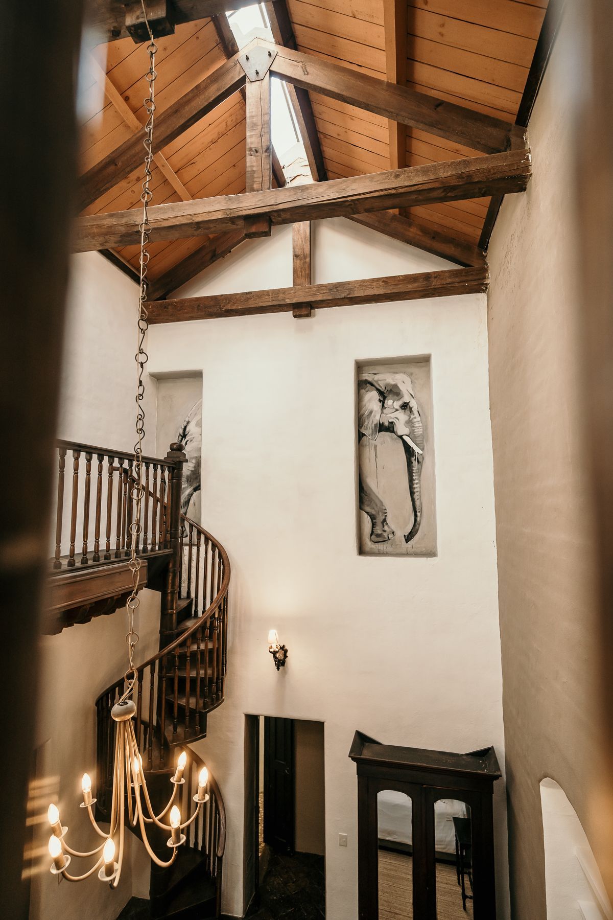Vista interior de una escalera y entrada con vigas de madera, una lámpara de araña y obras de arte.