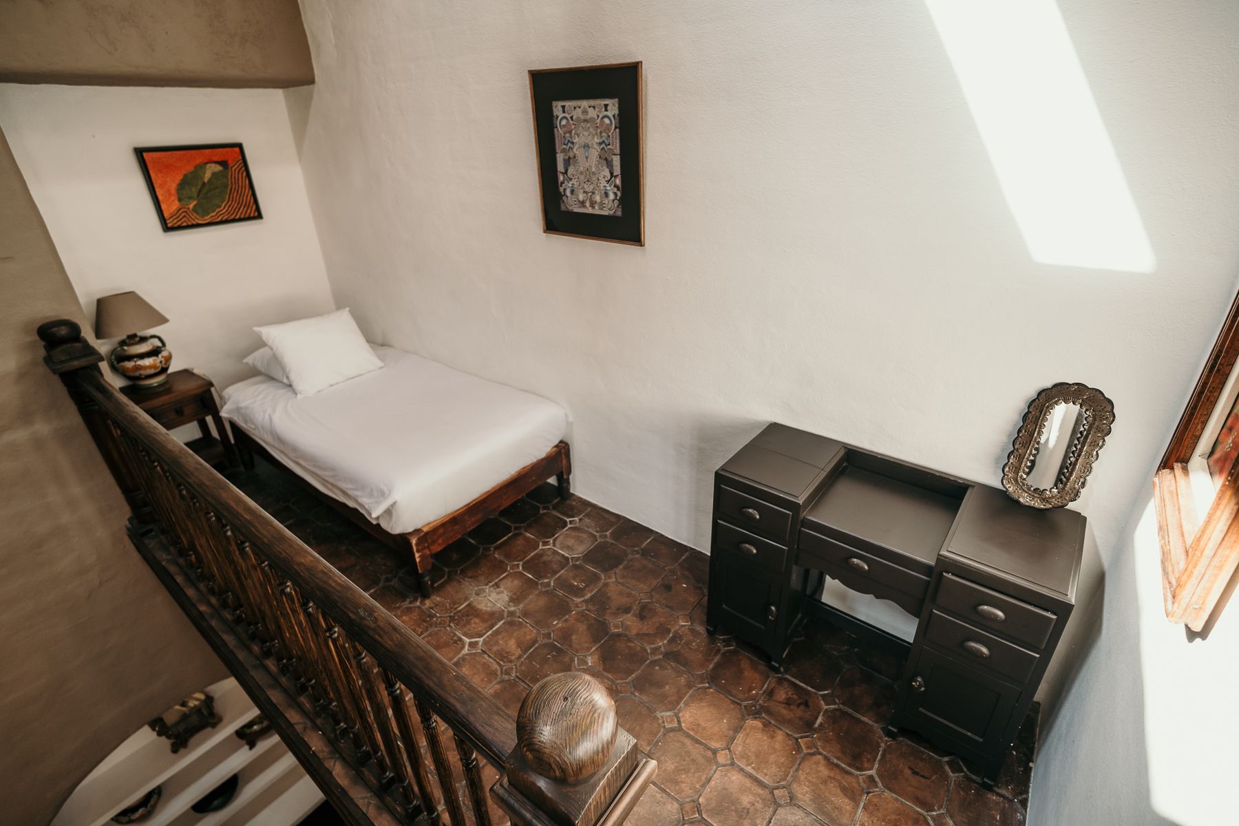 Zona de dormitorio con cama, escritorio y obras de arte sobre suelo de baldosas.