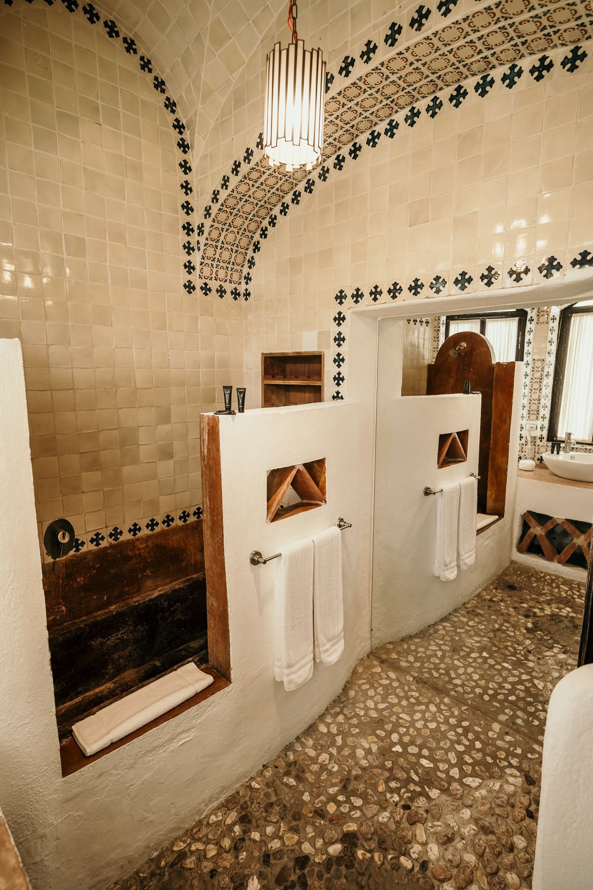 Baño con paredes de azulejos, suelo de piedra y techo abovedado. Dos duchas y lavabo.