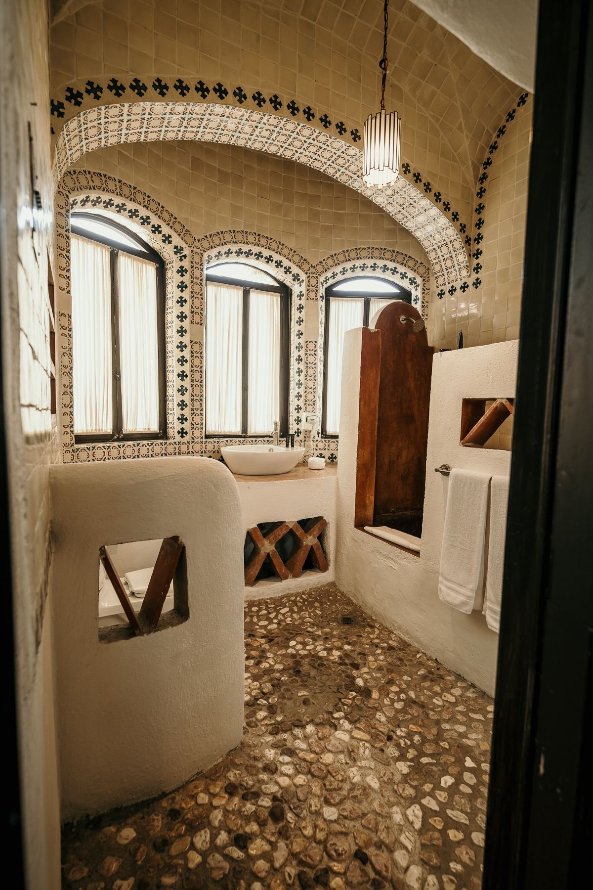 Baño con ventanas arqueadas, suelo de guijarros y paredes blancas con azulejos decorativos.