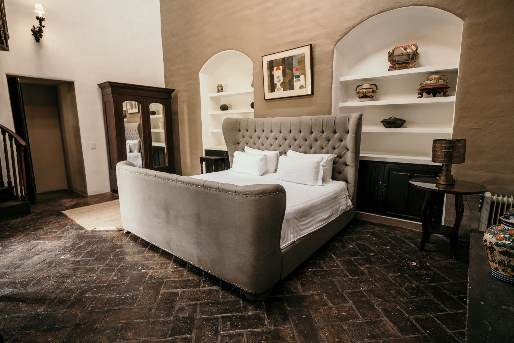 Dormitorio con una gran cama gris, estantes arqueados, muebles de madera oscura y piso de ladrillo.