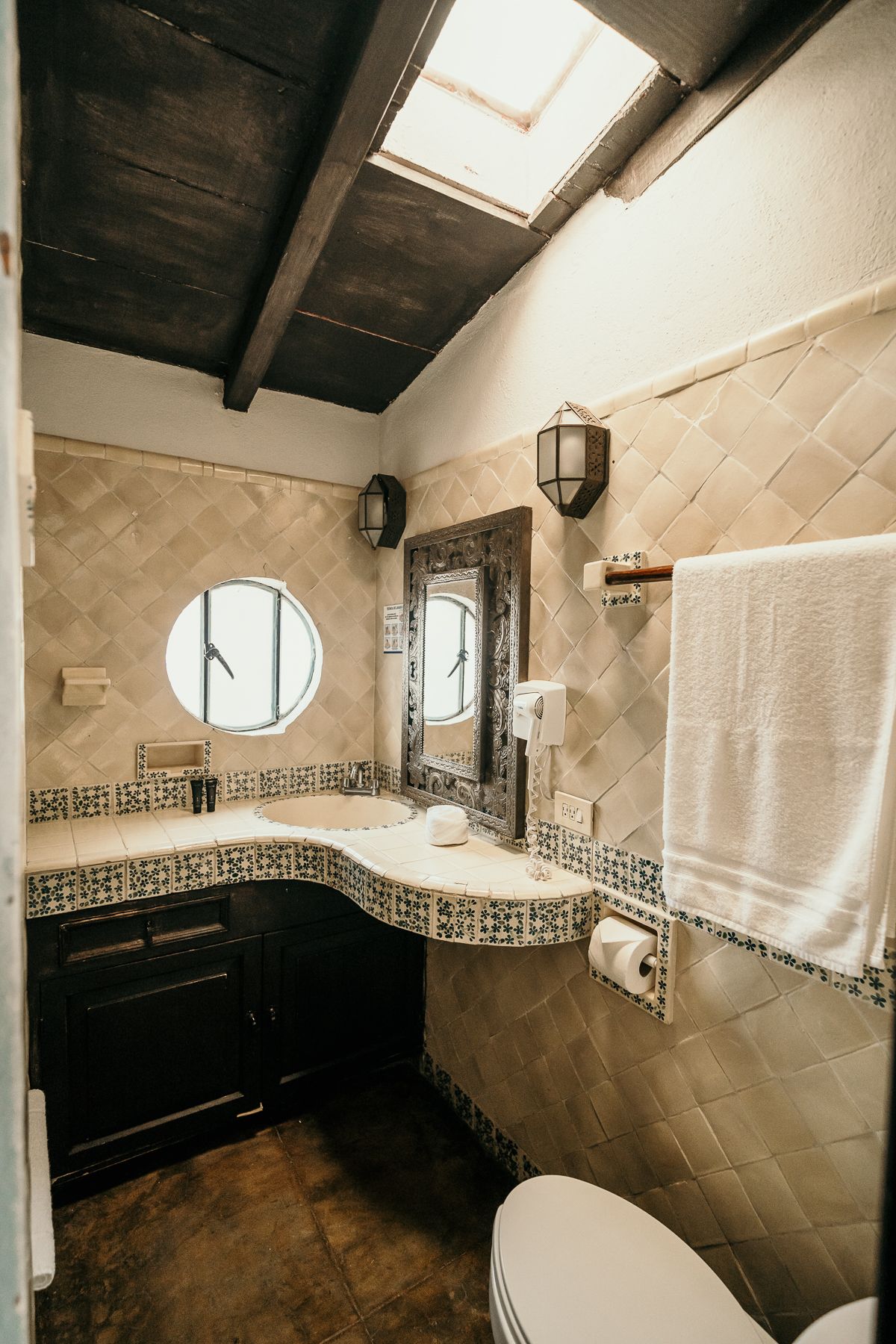 Baño con paredes de azulejos, mueble de madera oscura, ventana ovalada y tragaluz.