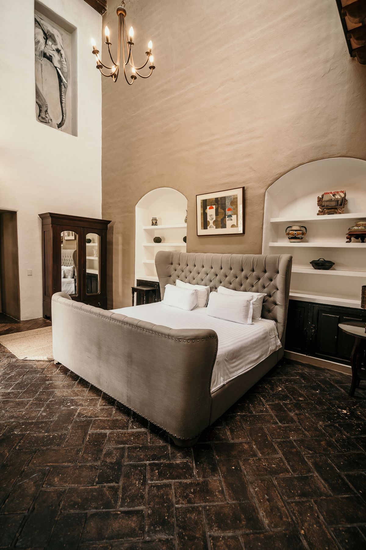 Dormitorio con cama grande, cabecera capitoné, estantes arqueados, piso de ladrillo, lámpara de araña y ventana alta.