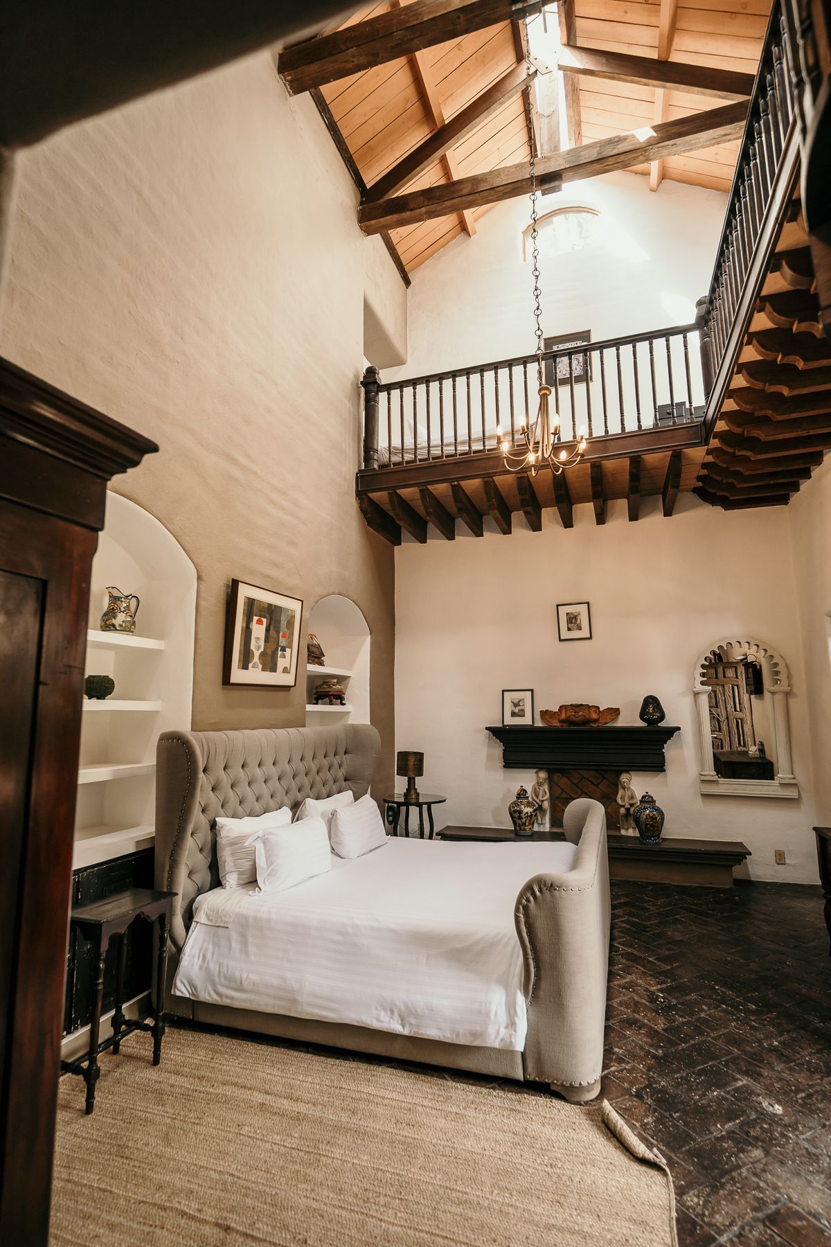 Dormitorio con techo alto, balcón, cama grande, chimenea, vigas de madera, tonos neutros.