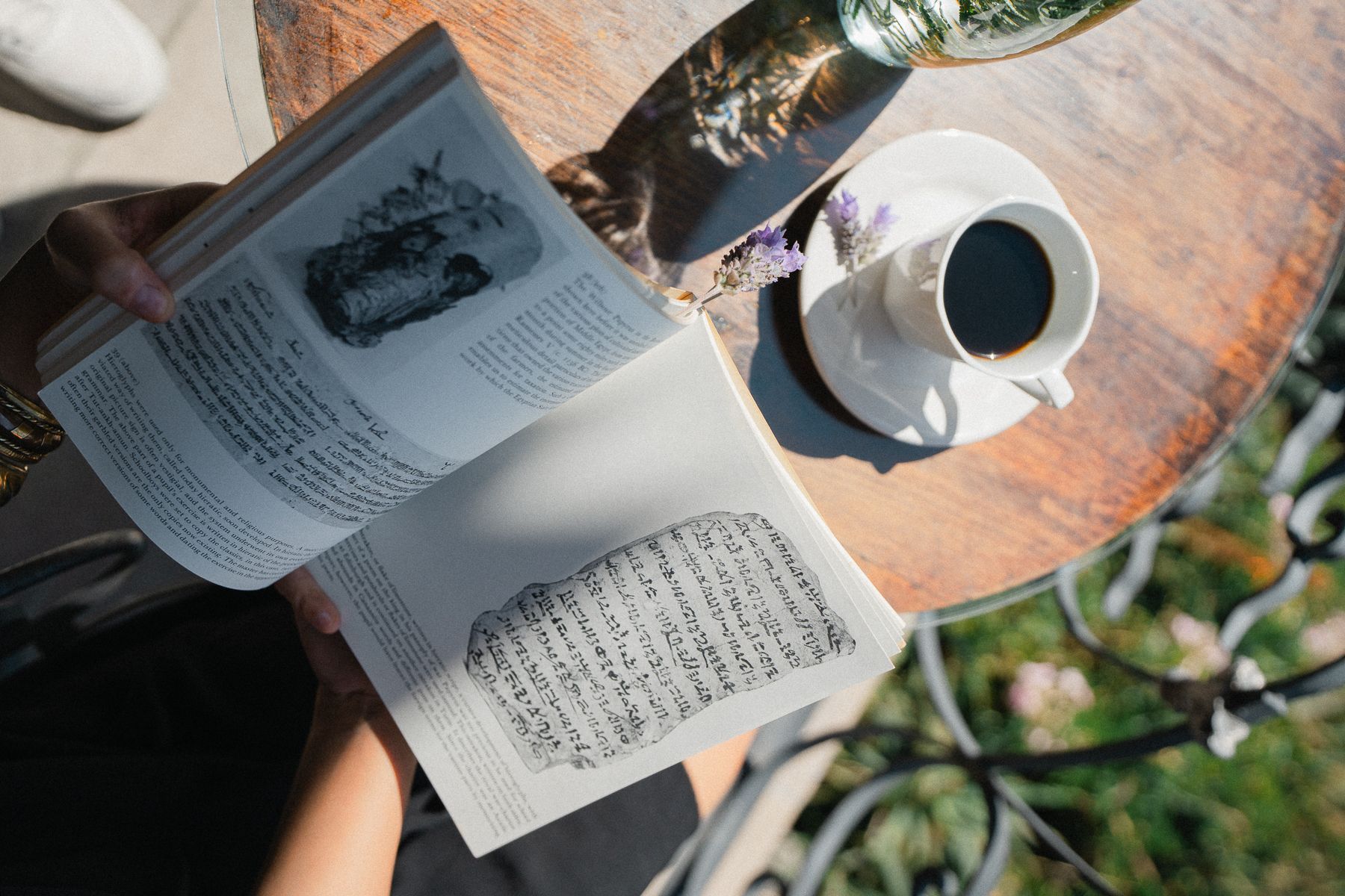 Persona leyendo un libro en una mesa con una taza de café. La luz del sol proyecta sombras.
