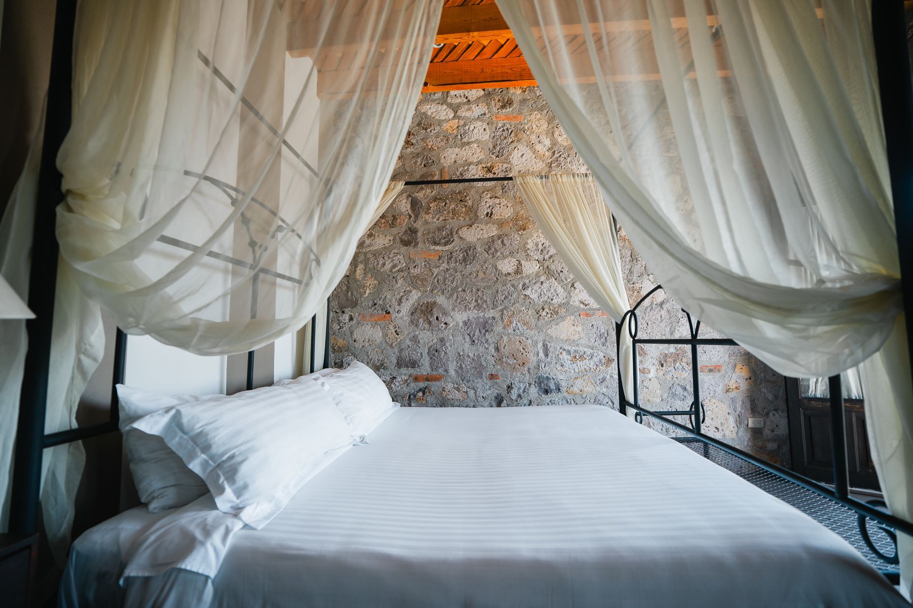 Cama con sábanas blancas y cortinas transparentes frente a una pared de piedra.