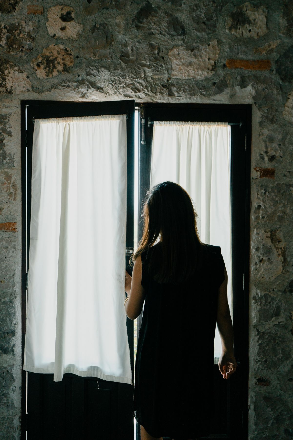 Mujer con vestido oscuro de pie en una puerta con cortinas blancas, luz natural.