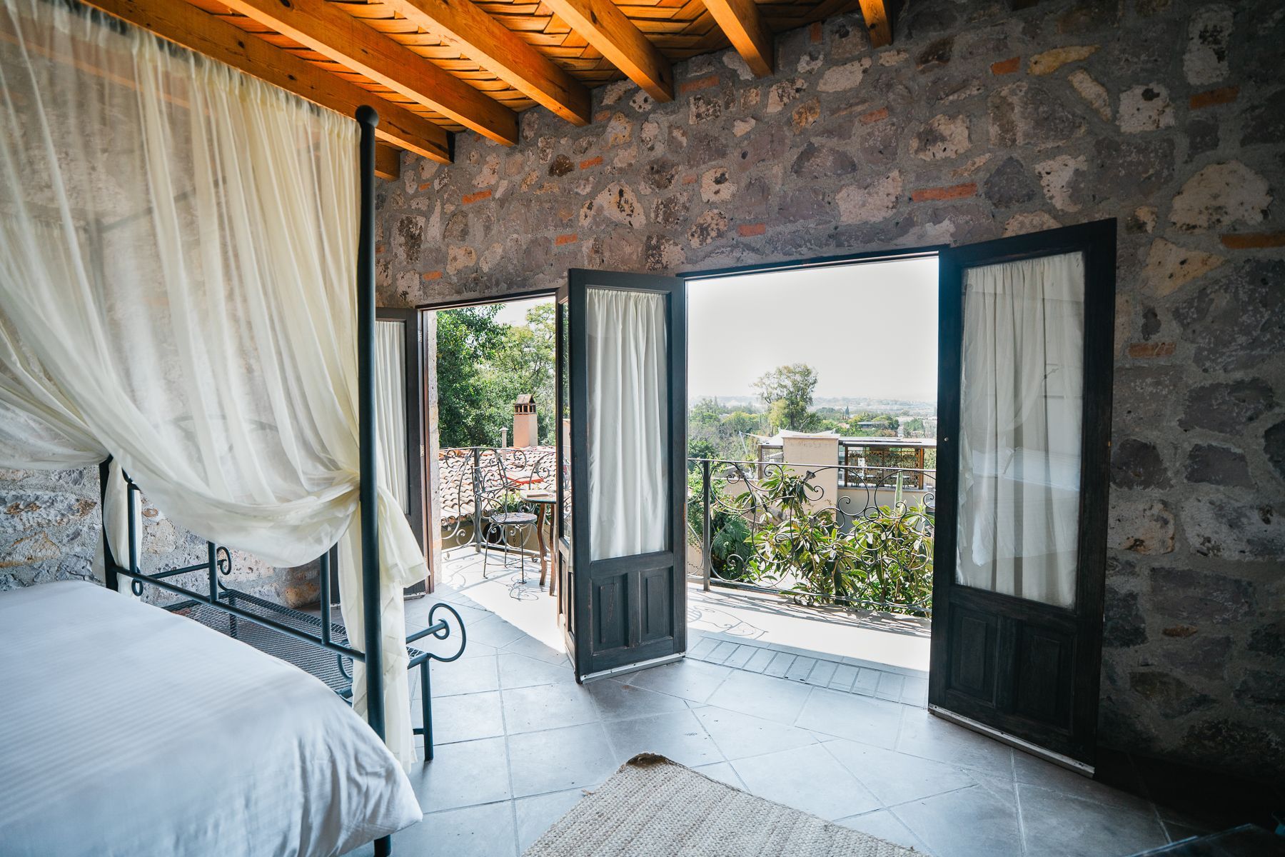 Dormitorio con paredes de piedra y puertas abiertas a un patio. Sábanas blancas, cortinas transparentes y techo de madera.
