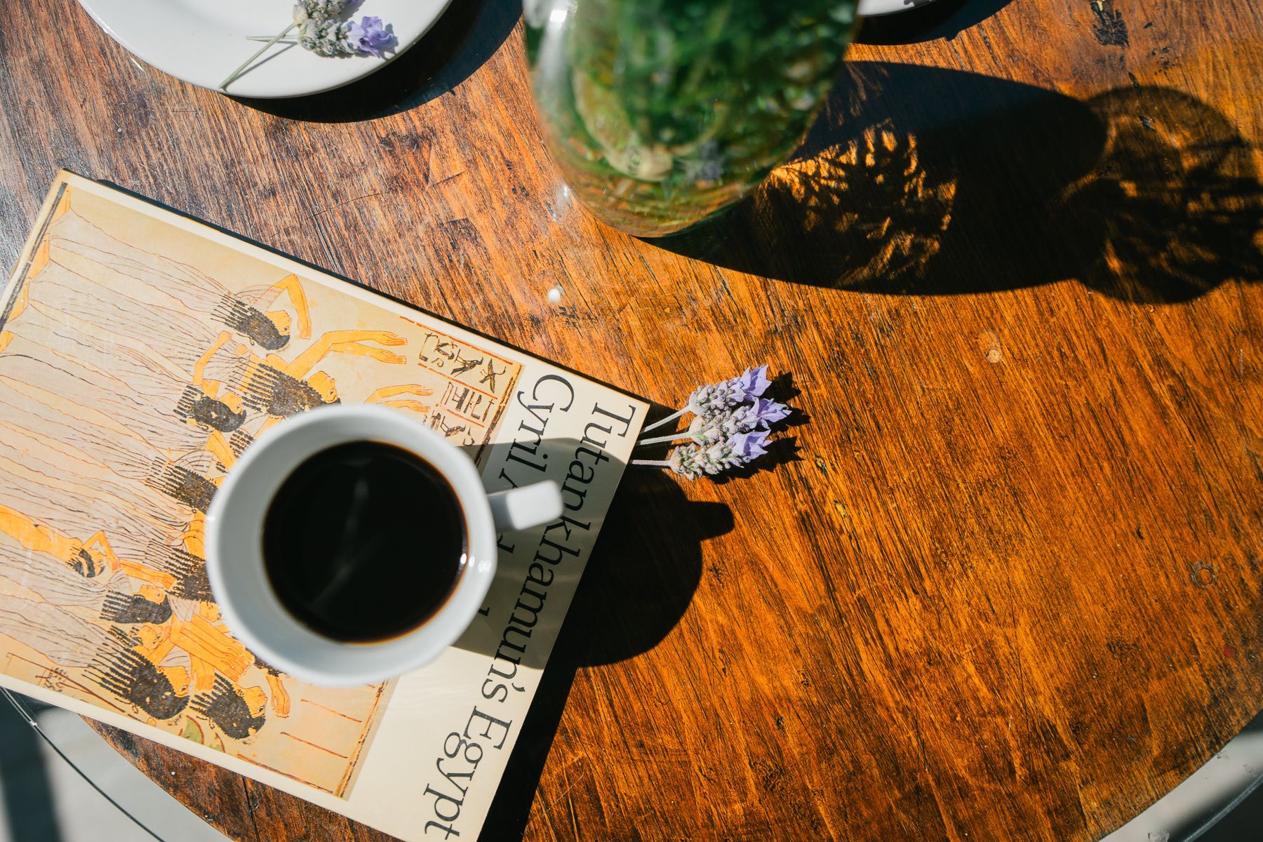 Taza de café sobre libro, lavanda y jarrón sobre una mesa de madera iluminada por el sol.