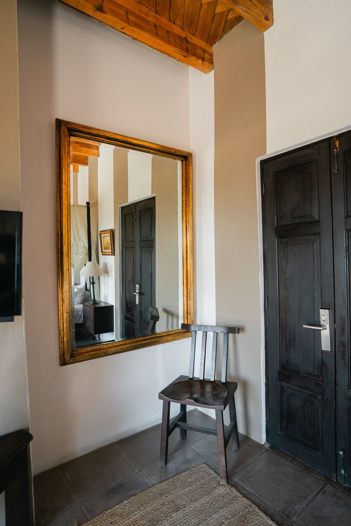 Pasillo con gran espejo con marco dorado, puertas de madera oscura y una silla de madera sobre una alfombra texturizada.