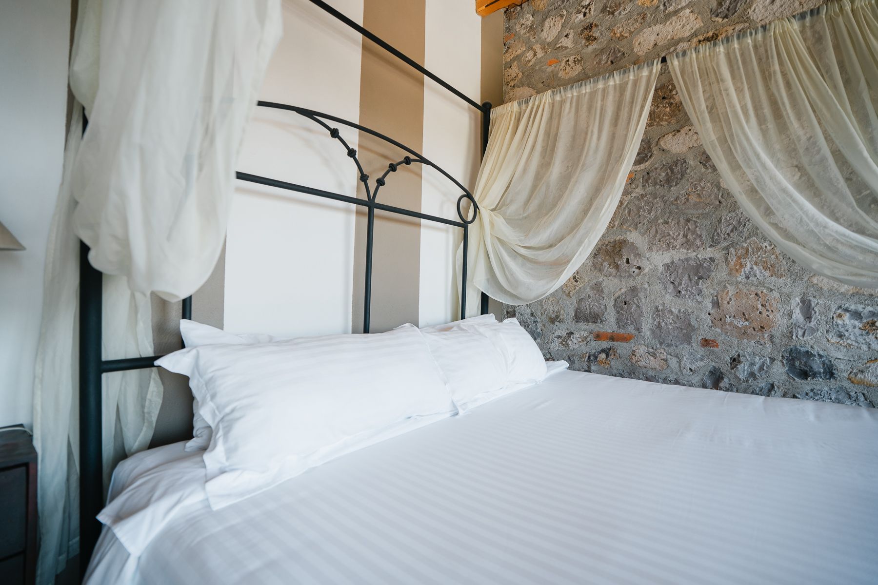 Cama blanca con dosel contra pared de piedra, pared de rayas beige y blanca detrás del cabecero.