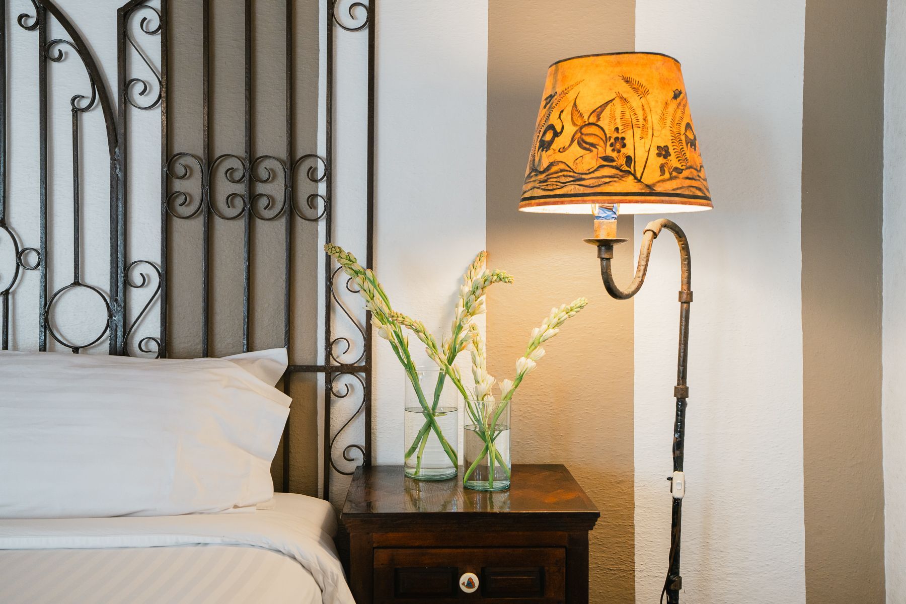 Escena de dormitorio: cama blanca, mesita de noche oscura con flores, lámpara de pie adornada con pantalla estampada.