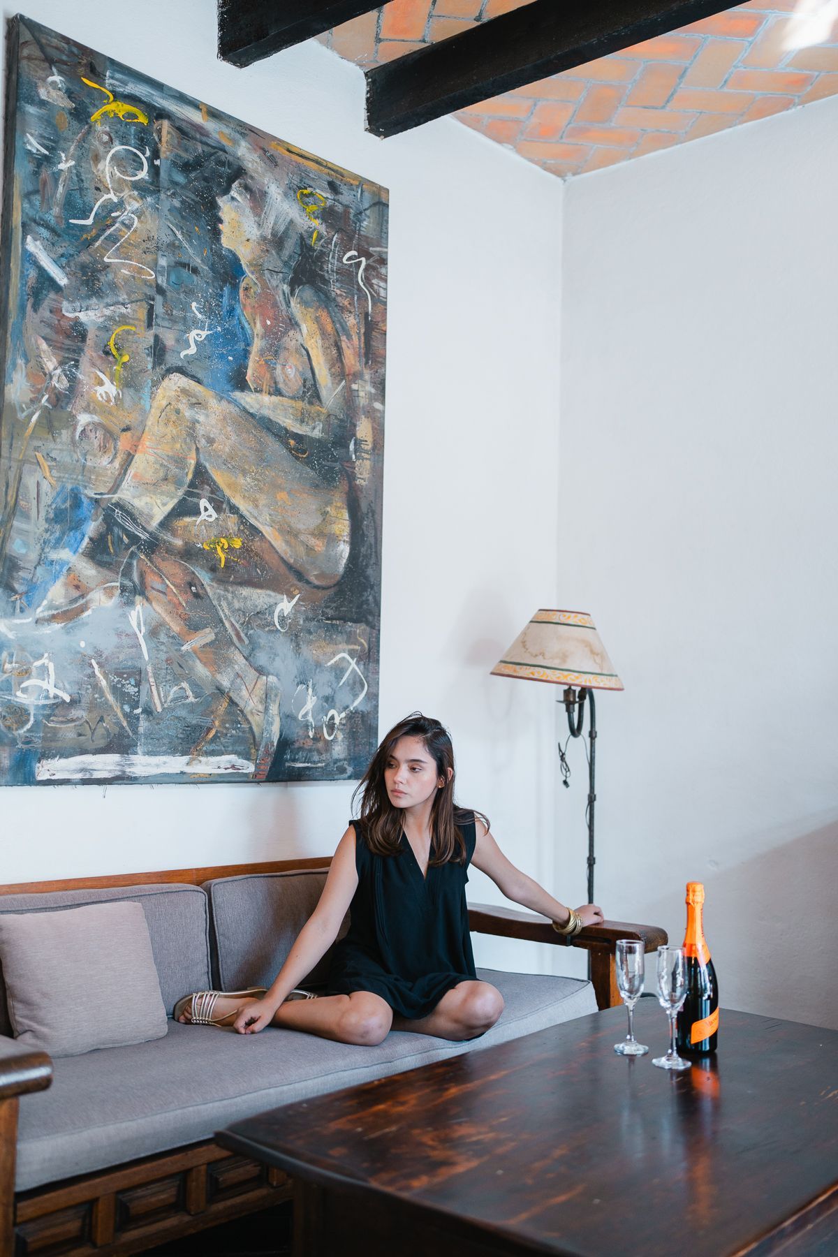 Una mujer se sienta en un sofá junto a una mesa con champán y copas, cerca de un gran cuadro abstracto.