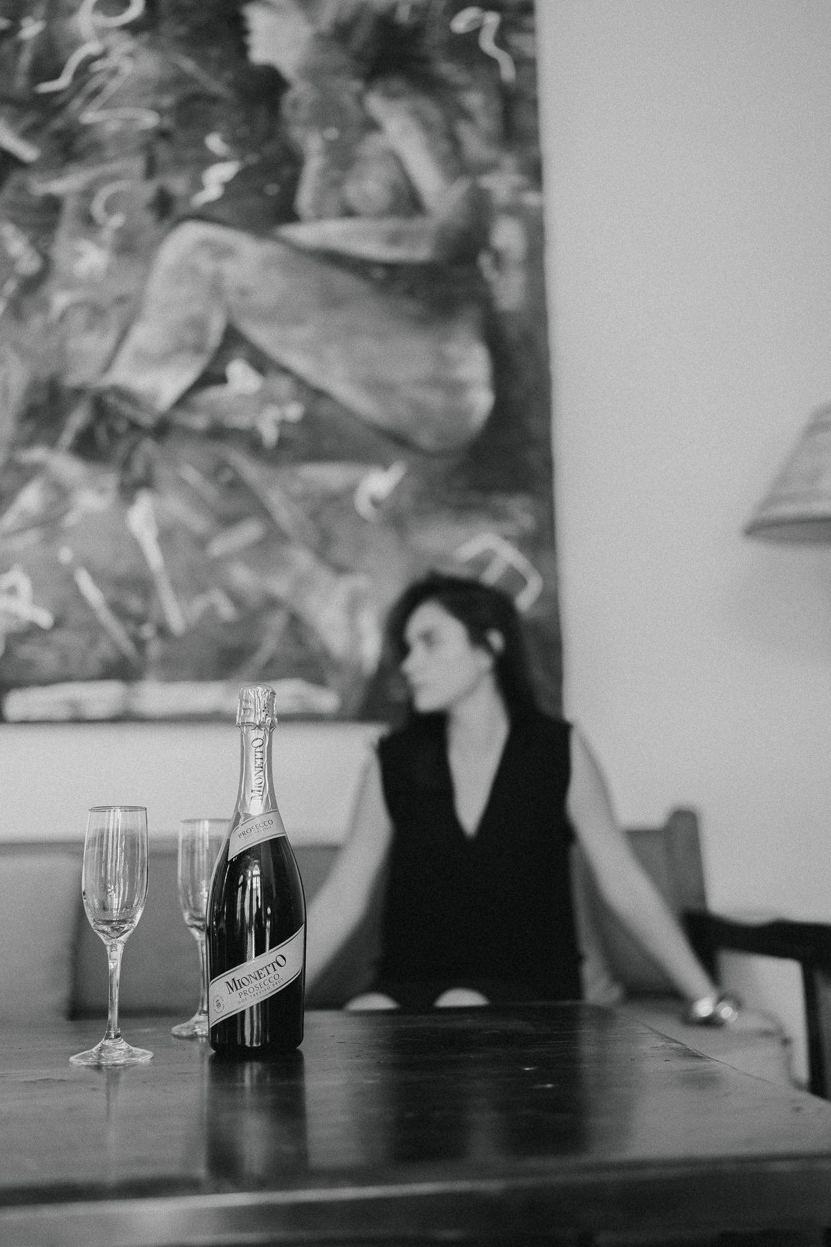 Mujer sentada en una mesa con champán y copas; hay un cuadro al fondo.