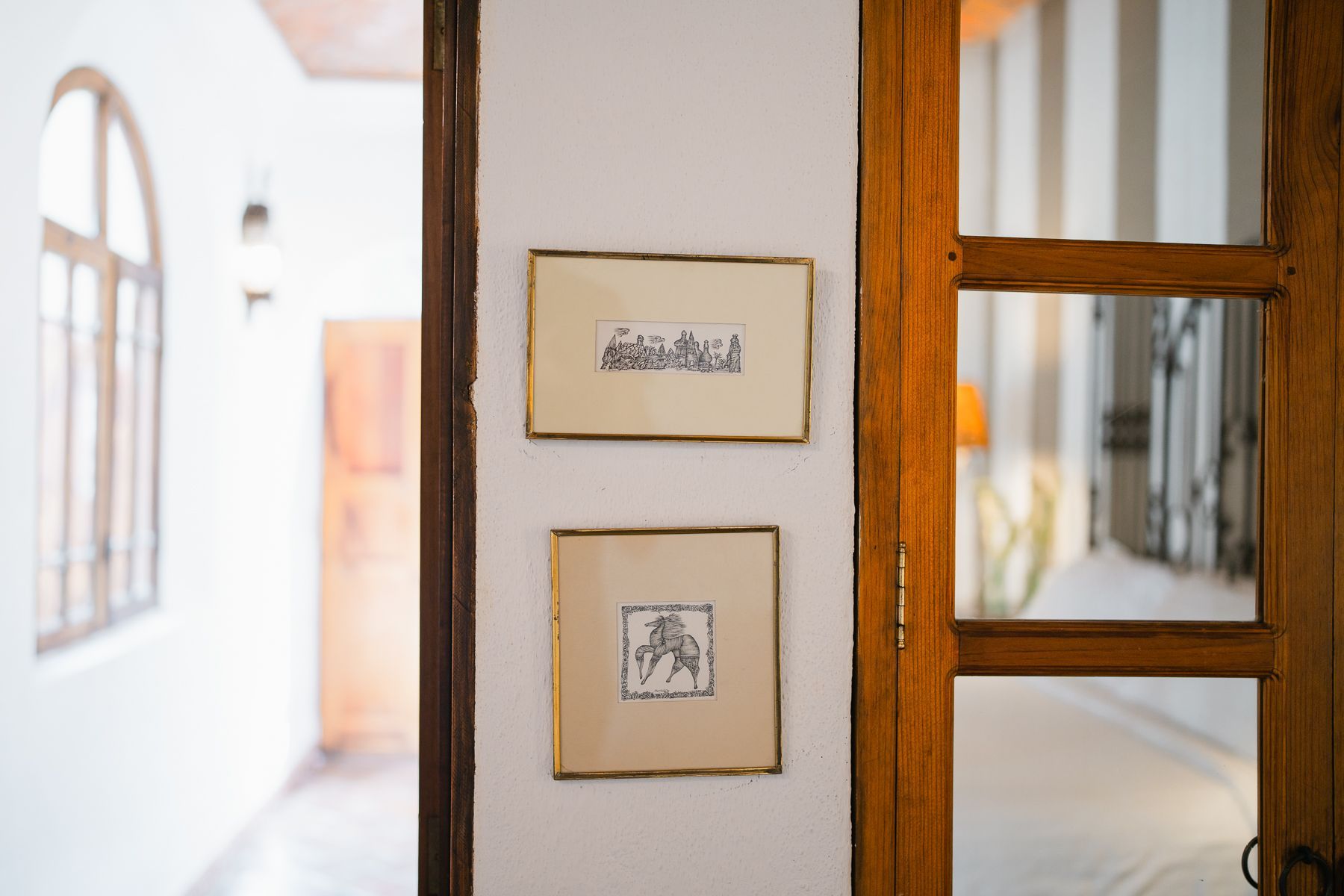 Dos dibujos enmarcados en una pared blanca; marco de puerta de madera a la derecha; ventana arqueada a la izquierda.