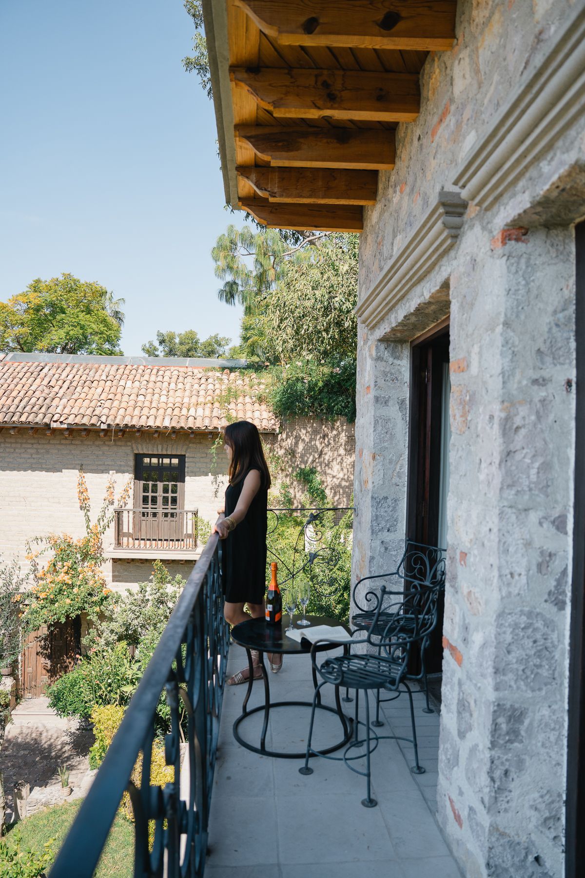 Mujer en un balcón mirando hacia afuera. Edificio de piedra, sillas de hierro forjado y vegetación al fondo. Día soleado.