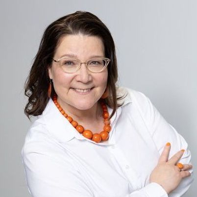 Päivi Saatsi wearing glasses and an orange necklace is smiling for the camera.