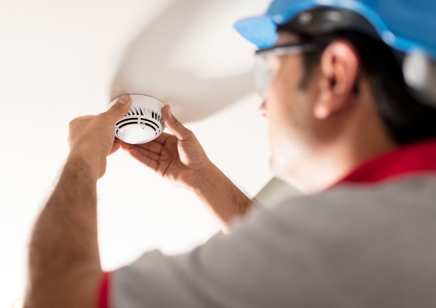 maintenance worker in a blue hardhat inspecting a smoke detector