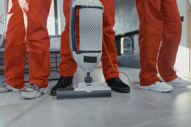 three professional cleaners in orange overalls standing side-by-side, middle person holding a white vacuum