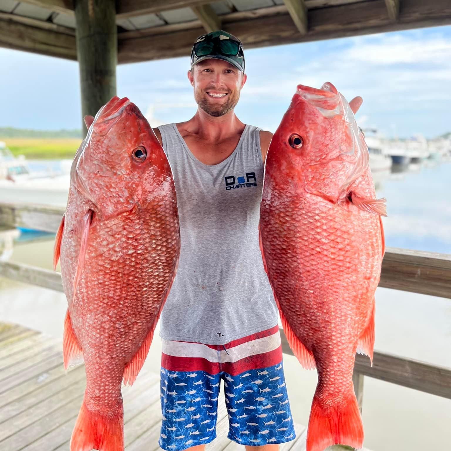 A man is holding two large red fish on a dock.