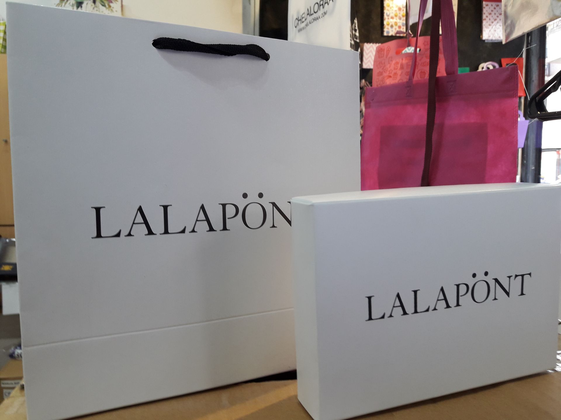 Sobre una mesa hay una bolsa de la compra blanca y una caja de regalo rectangular, ambas con el logotipo negro 