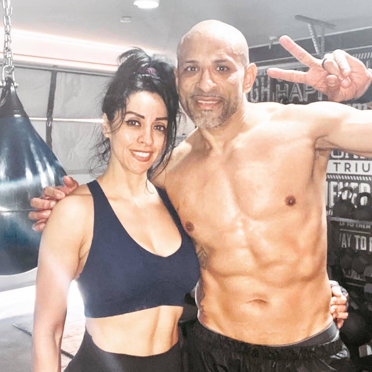 A smiling woman and a shirtless man with athletic builds posing for a photo in a gym with a boxing bag.