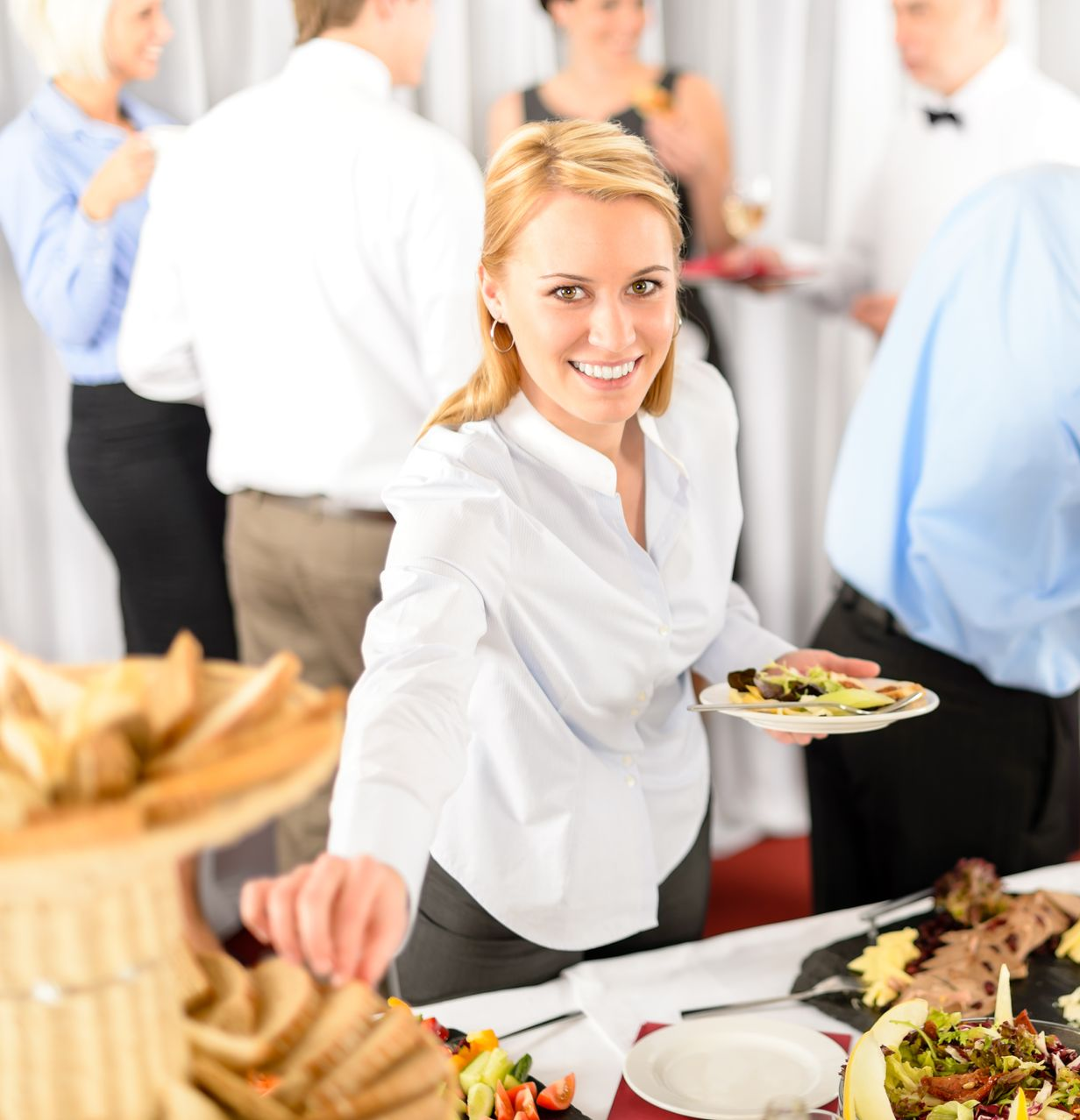 Eine Frau lächelt am Buffet und greift nach Essen. Im Hintergrund sind Menschen bei einer Veranstaltung zu sehen.