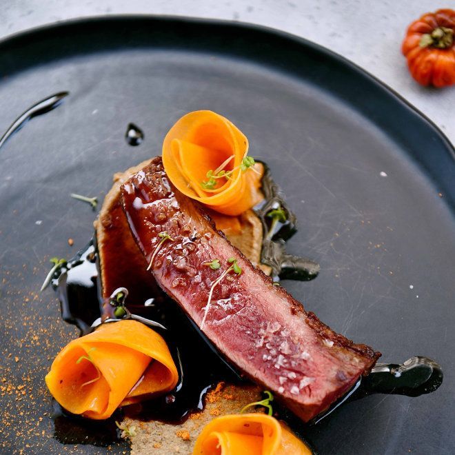 Gebratenes Steak mit Karottenblüten, Sauce und Kräutern auf einem schwarzen Teller.