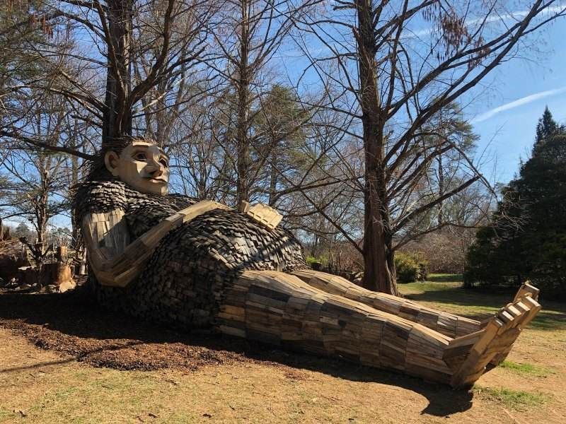 A large wooden statue of a woman laying on the ground in a park.