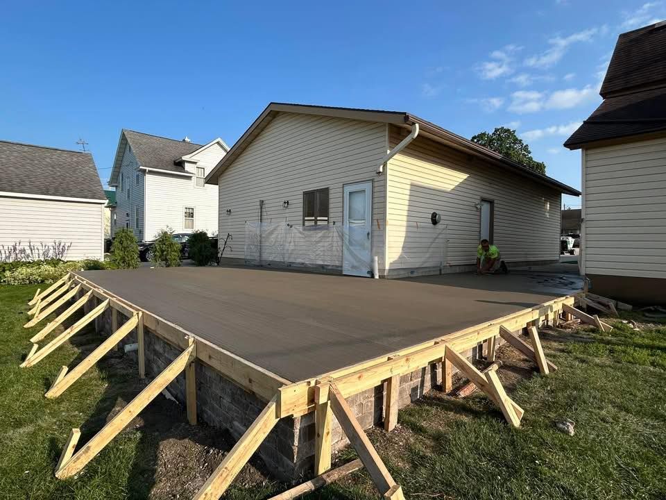 Newly poured concrete patio adjacent to a beige building, supported by wooden framing.