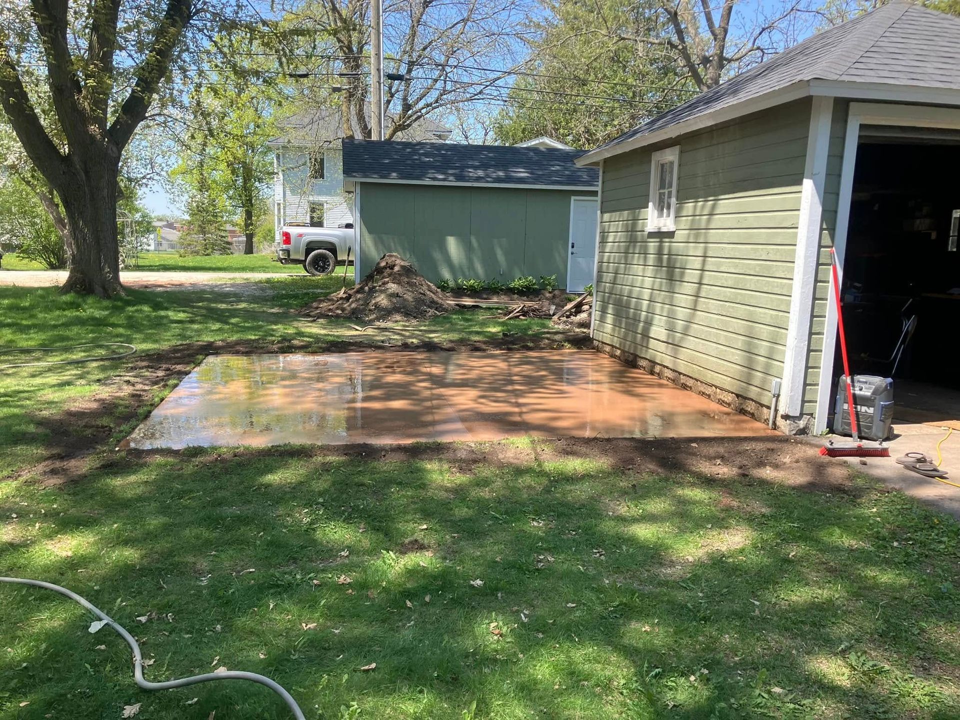 Concrete patio next to a green garage. Lawn in front with a pile of dirt.