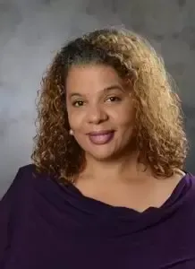 A woman with curly hair is wearing a purple shirt and smiling for the camera.