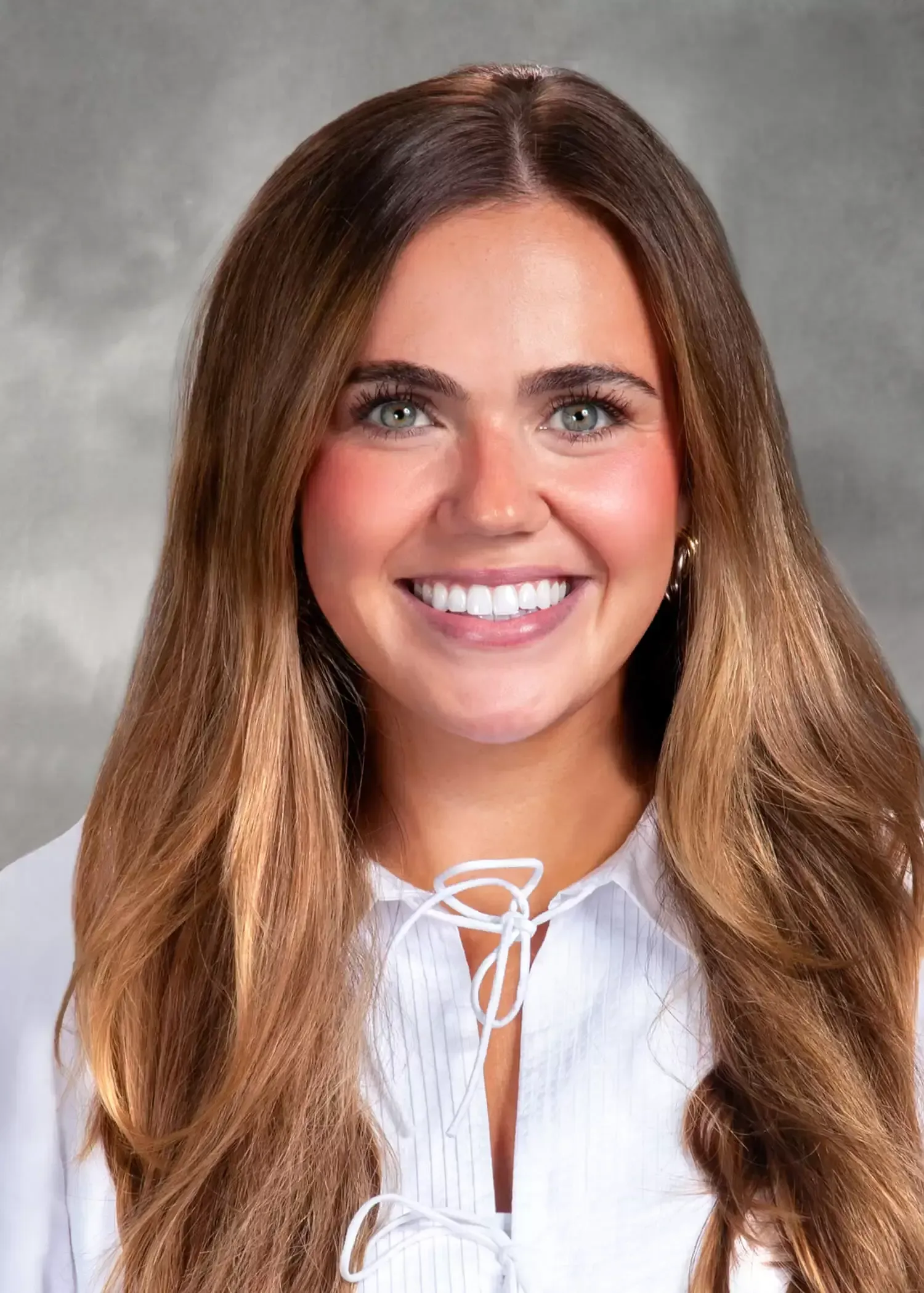 A woman with long brown hair is wearing a white shirt and smiling for the camera.