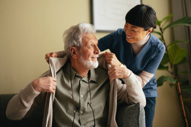 An elderly man is sitting on a couch while a nurse helps him get dressed.