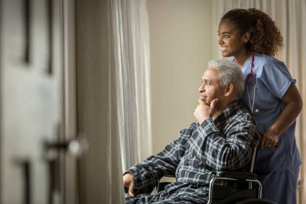 An elderly man in a wheelchair is looking out of a window with a nurse.