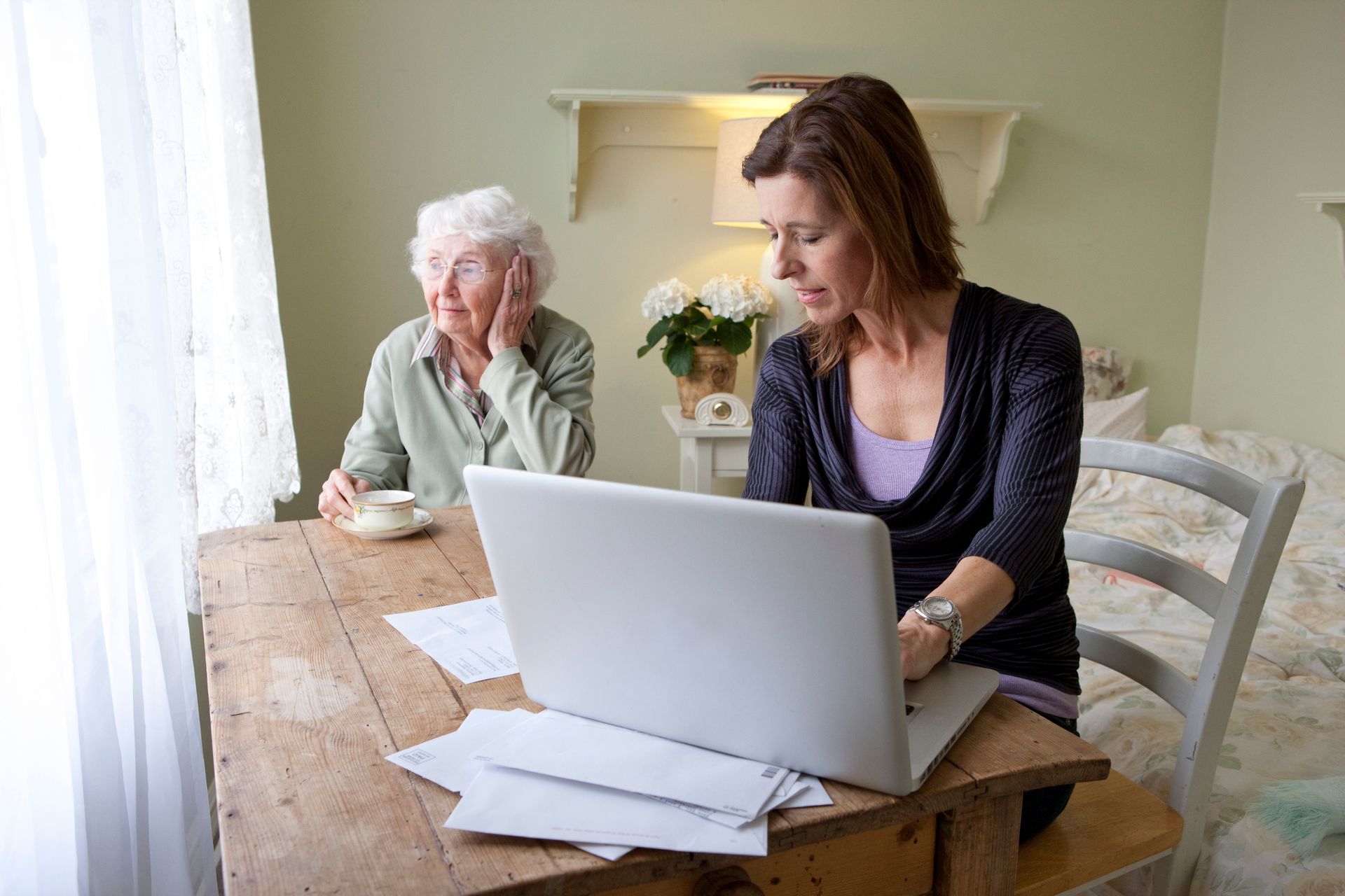 Caregiver and elderly woman at home using laptop, reviewing documents at wooden table