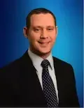 A man in a suit and tie is smiling in front of a blue background.