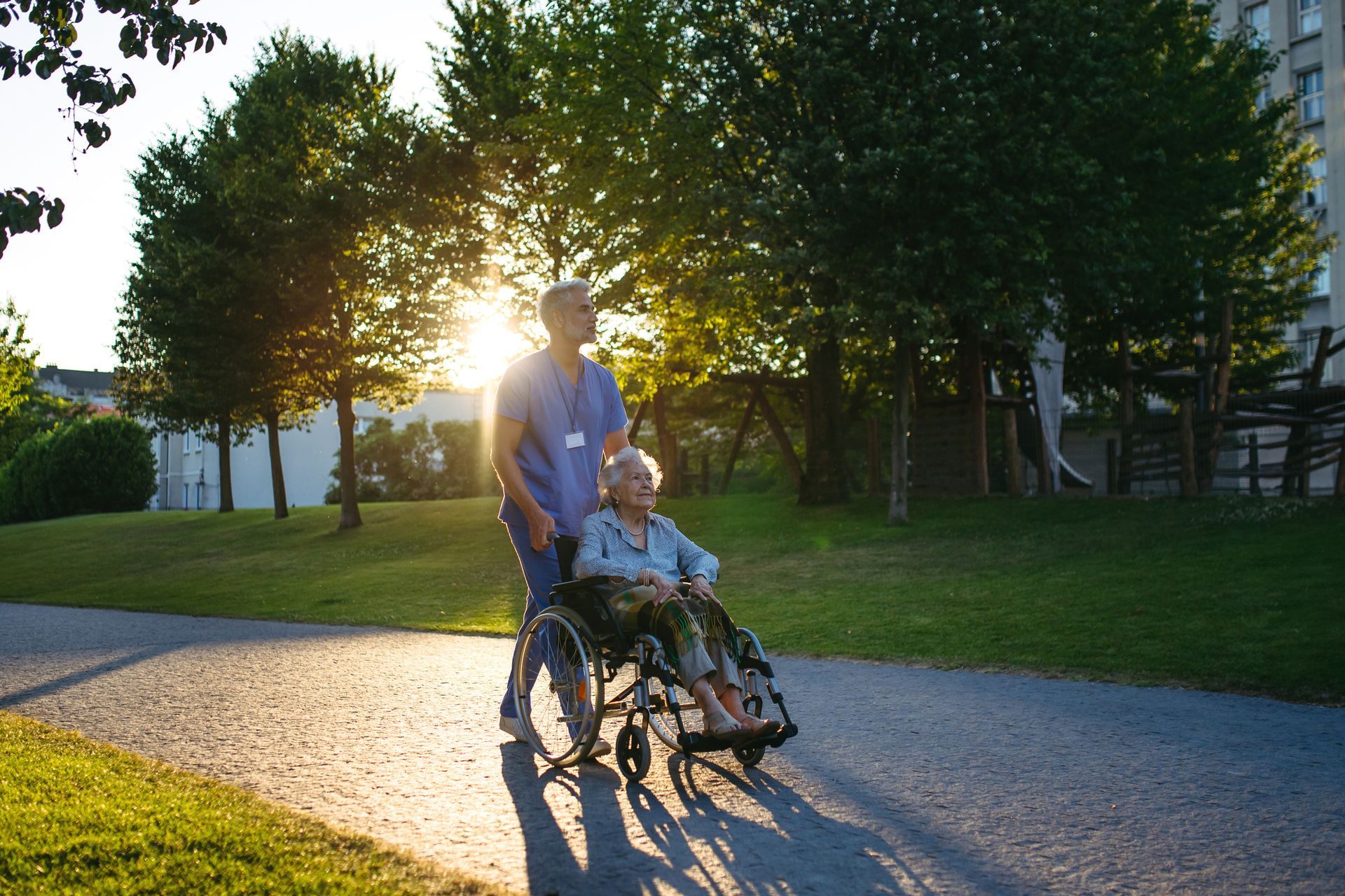 Caregiver pushing an elderly woman in a wheelchair along a sunny path, trees and a building in the background.
