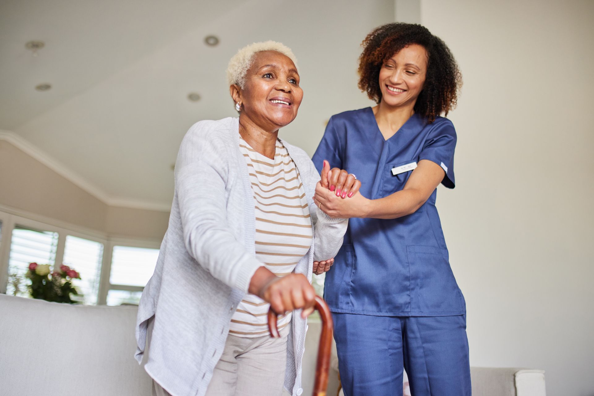 Caregiver assisting an older adult with a cane in a bright living room.