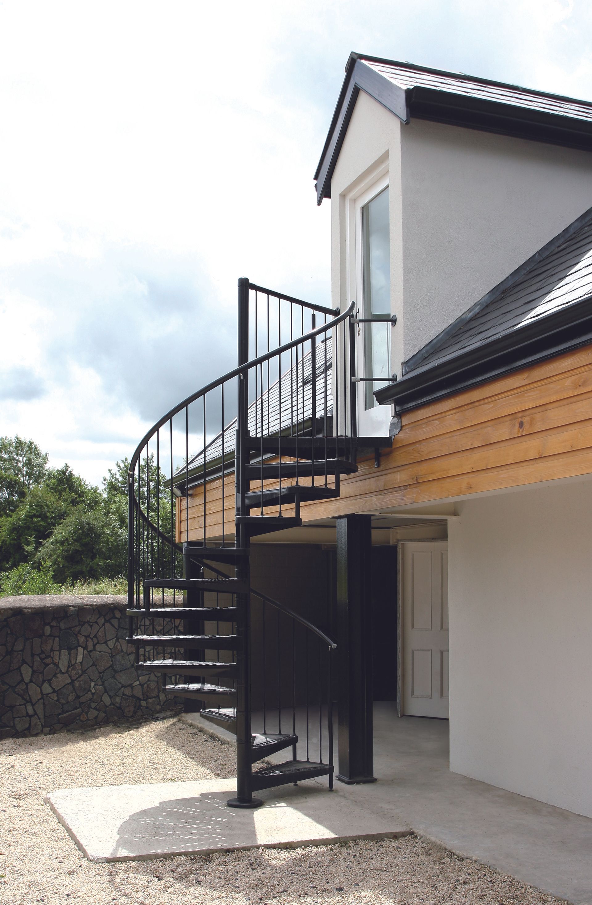Esterno di una casa con scala a chiocciola nera e balcone, pareti bianche e tegole scure sul tetto.