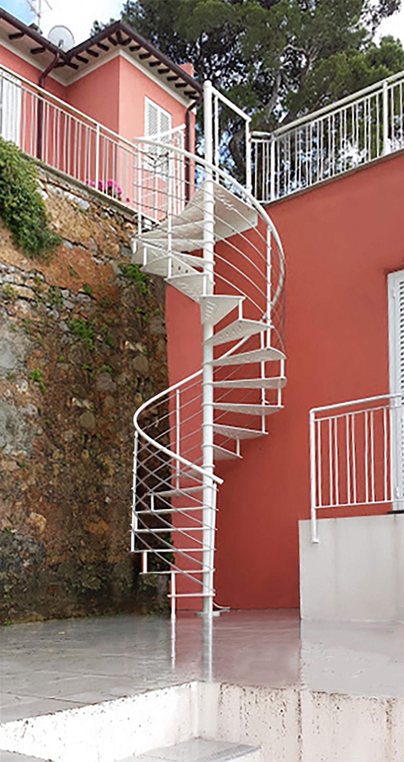Scala a chiocciola bianca accanto a un edificio color rosso corallo e a una parete rocciosa, con ringhiere bianche e pavimento lucido.