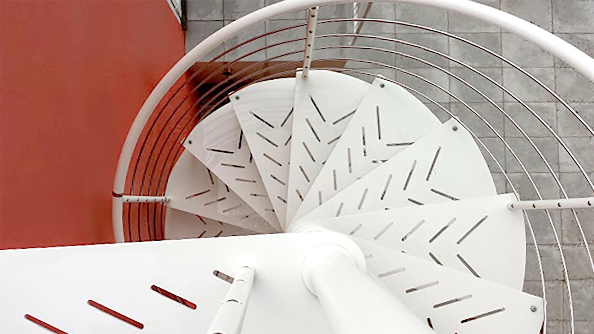 Scala a chiocciola bianca con ringhiere in metallo e gradini scanalati, addossata a una parete rossa.