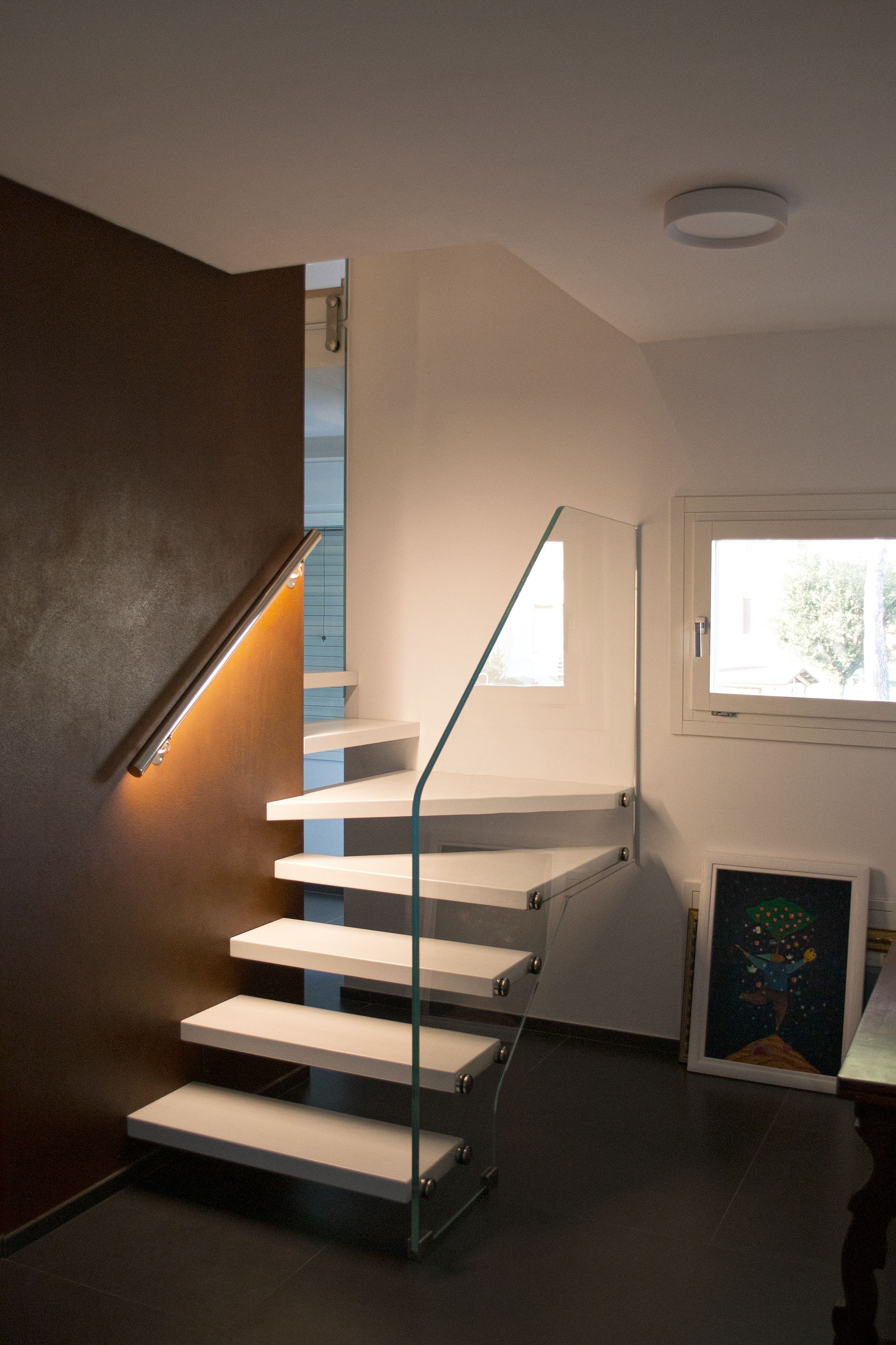 Scala interna moderna con gradini grigi, corrimano in legno e piccole finestre in un corridoio luminoso.