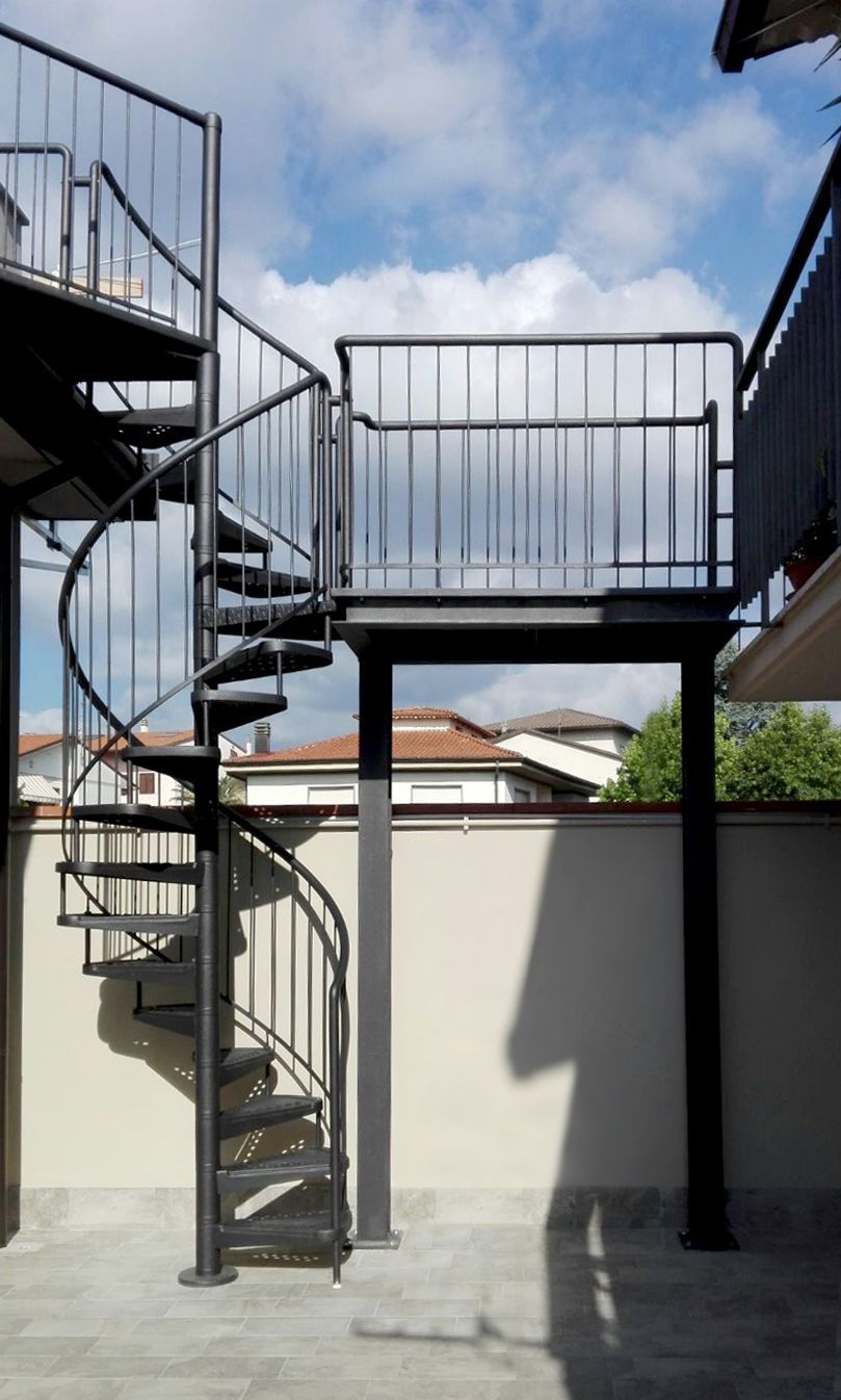 Scala a chiocciola in metallo e passerella sopraelevata si stagliano contro il muro bianco di un cortile sotto un cielo azzurro.