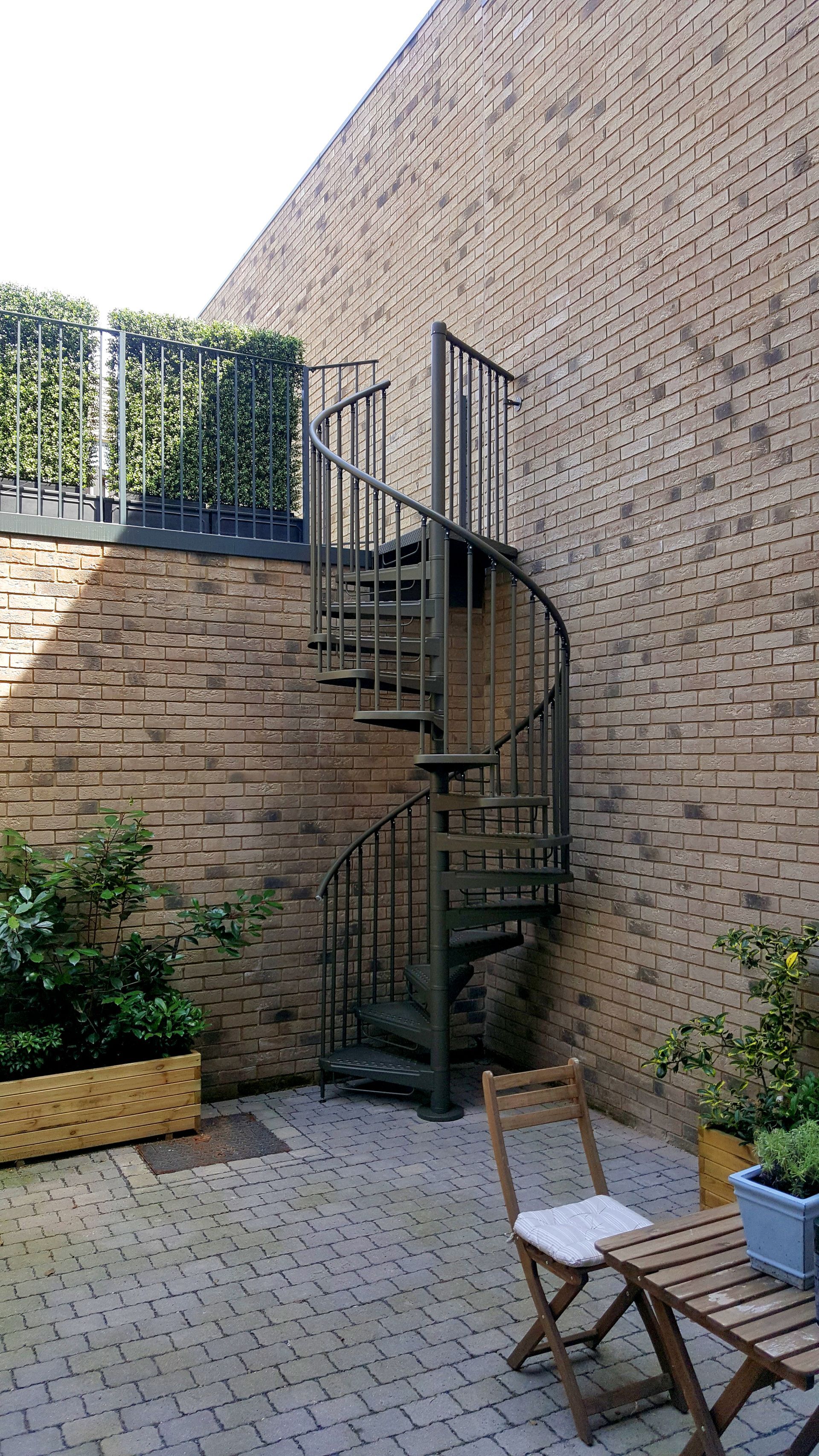 Cortile esterno in mattoni con scala a chiocciola in metallo nero e sedie da patio in primo piano.
