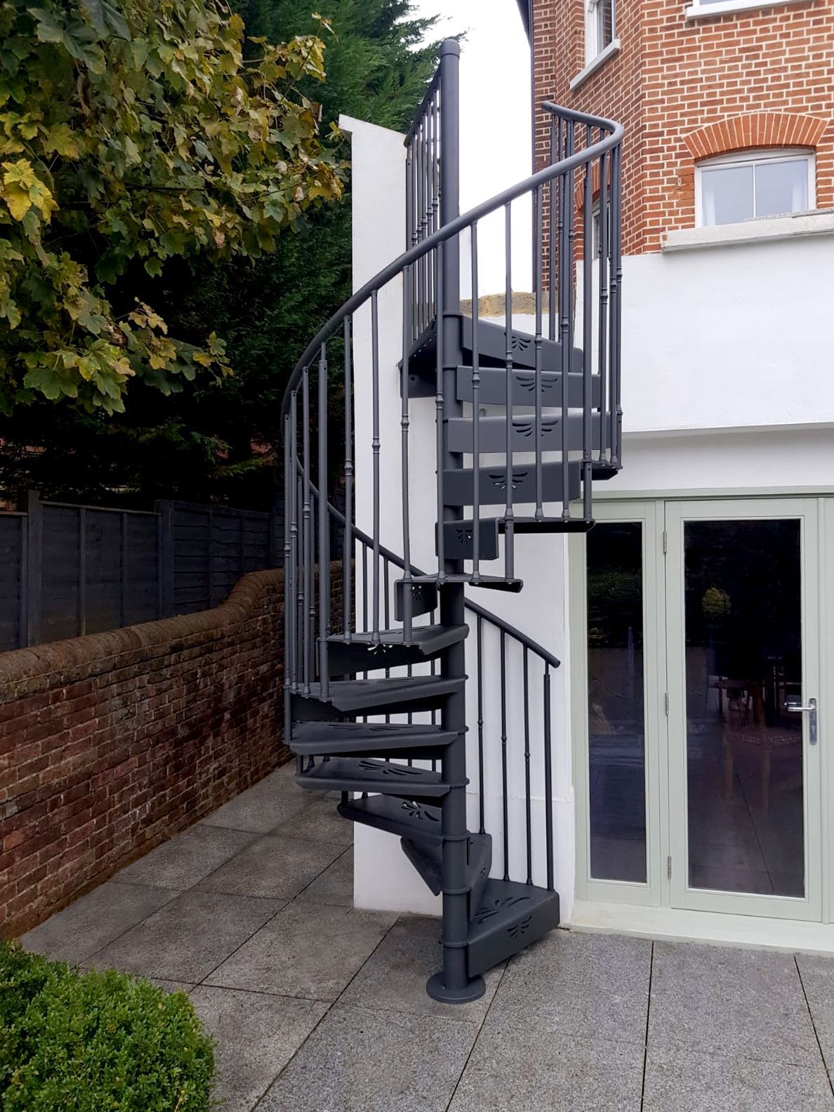 Scala a chiocciola in metallo nero accanto a una casa di mattoni bianchi con porte a vetri e un muro di mattoni.